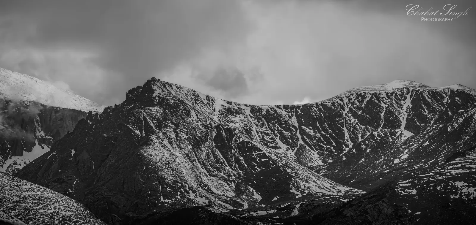 A rugged, snow-dusted mountain ridge beneath heavy storm clouds, in black and white.
