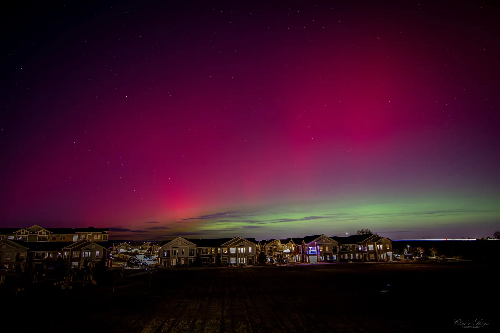 Vivid pink and green aurora borealis glowing over a row of houses under a starry sky.