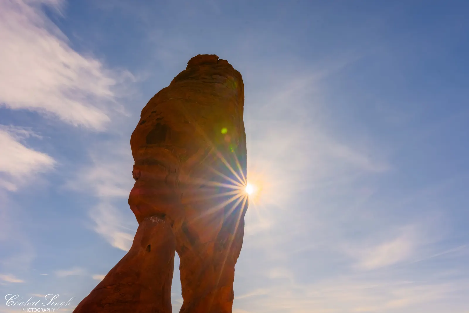 Delicate Arch silhouetted against a bright sunburst breaking through the blue sky.