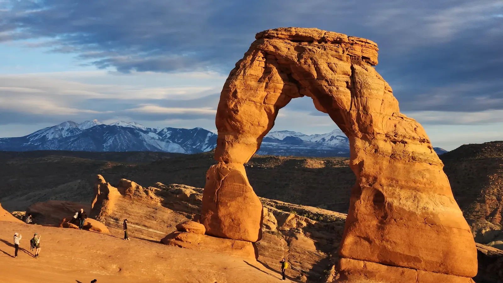 Delicate Arch glowing gold in late-afternoon light, snow-capped mountains in the distance.