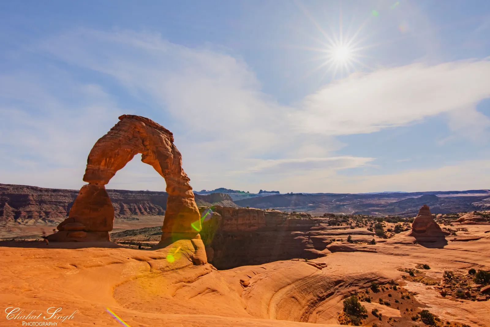 Delicate Arch with the sun peeking above the horizon, lens flare and desert mesas beyond.
