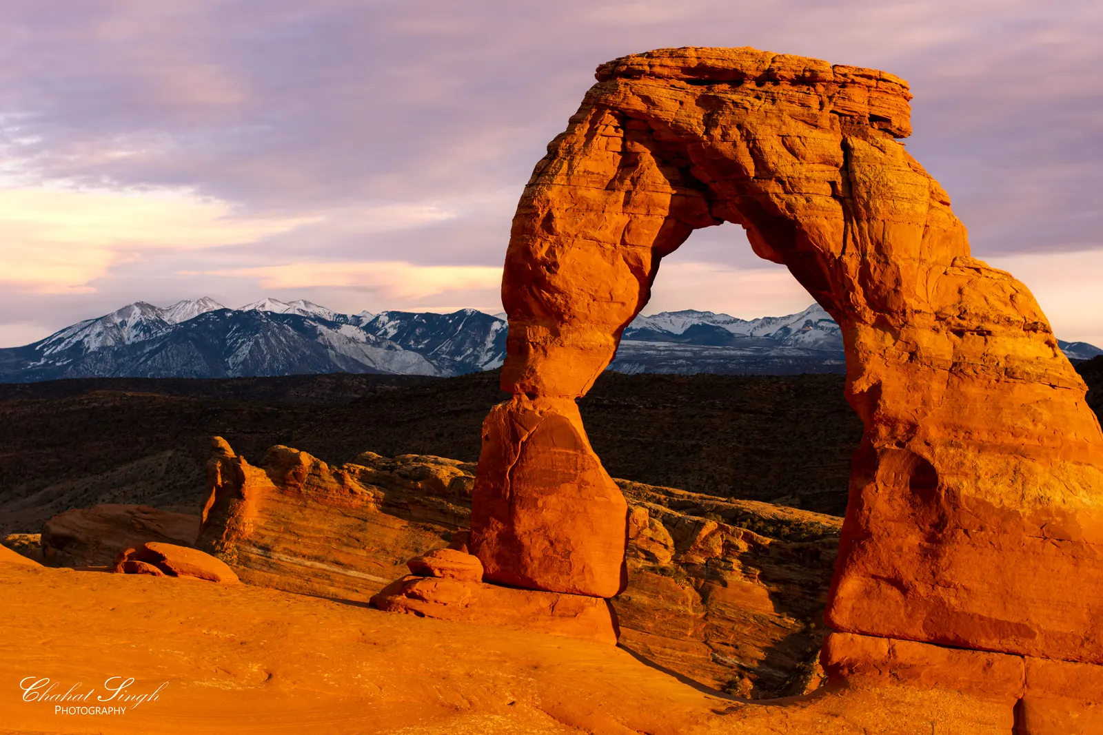 Delicate Arch glowing orange at sunset, snow-capped mountains and desert layers behind it.