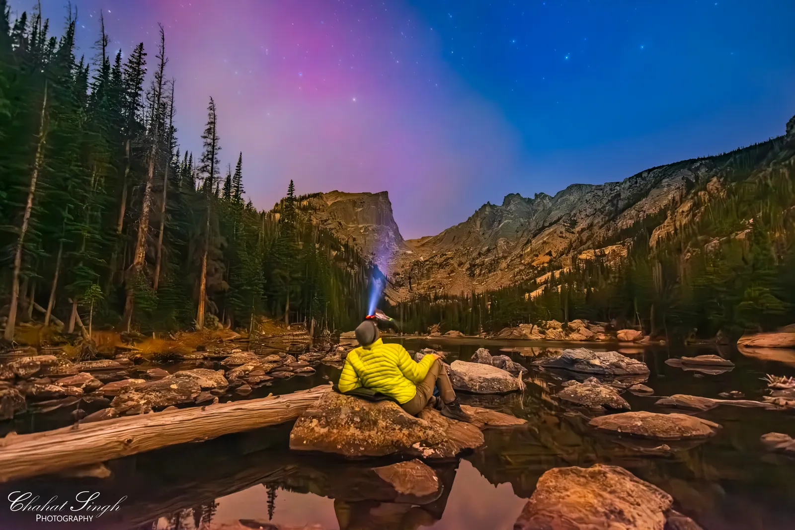 A person sitting on rocks beside Dream Lake, gazing up at a star-filled, aurora-tinted sky.