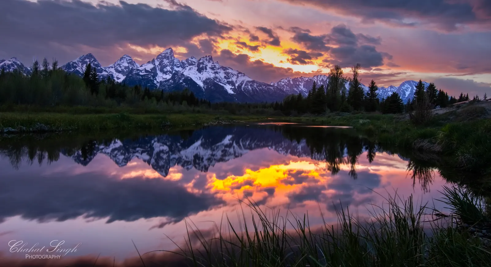 Grand Teton mountains under a fiery sunset sky, perfectly reflected in a calm pond.