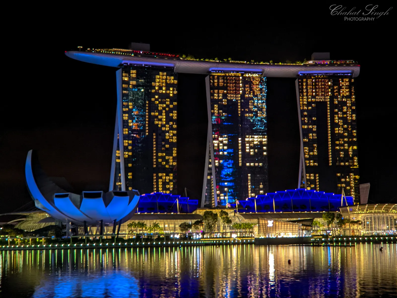 Marina Bay Sands hotel and its rooftop illuminated blue at night, reflected in the water.