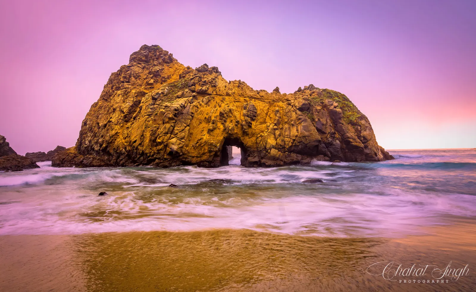 A sea arch at Pfeiffer Beach glowing gold at sunset, waves washing onto purple sand.