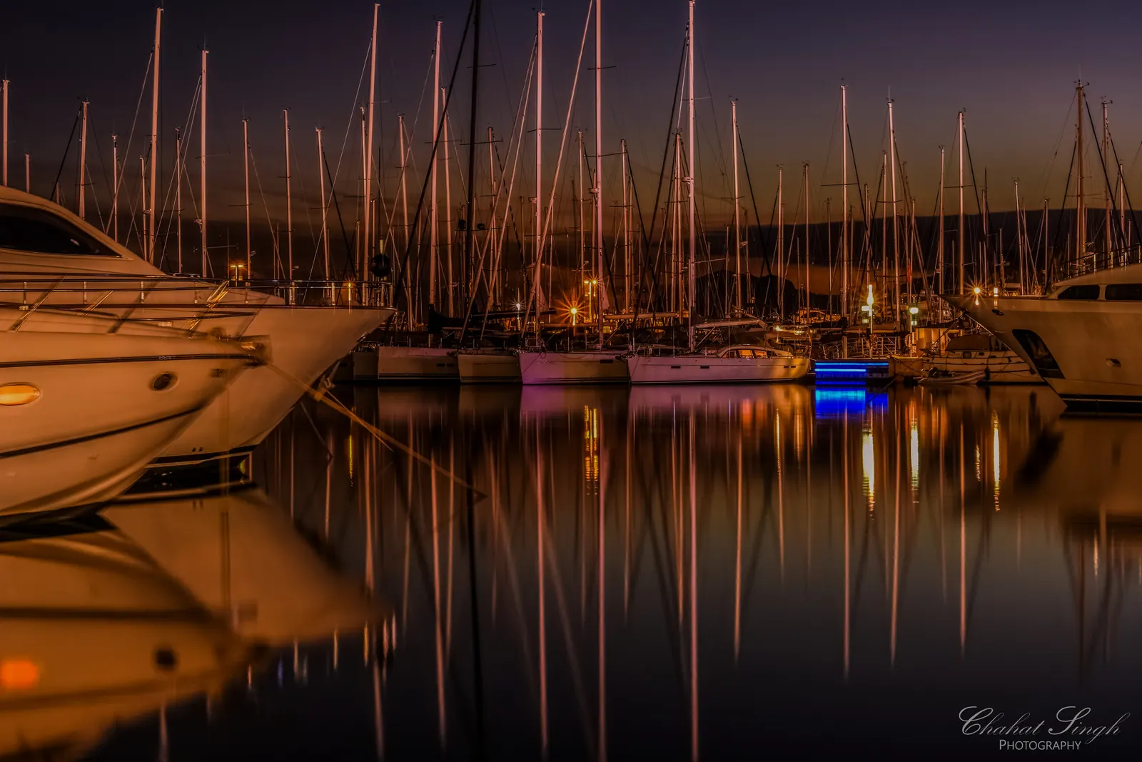 Sailboats and their masts reflected in still water at a marina during blue hour.