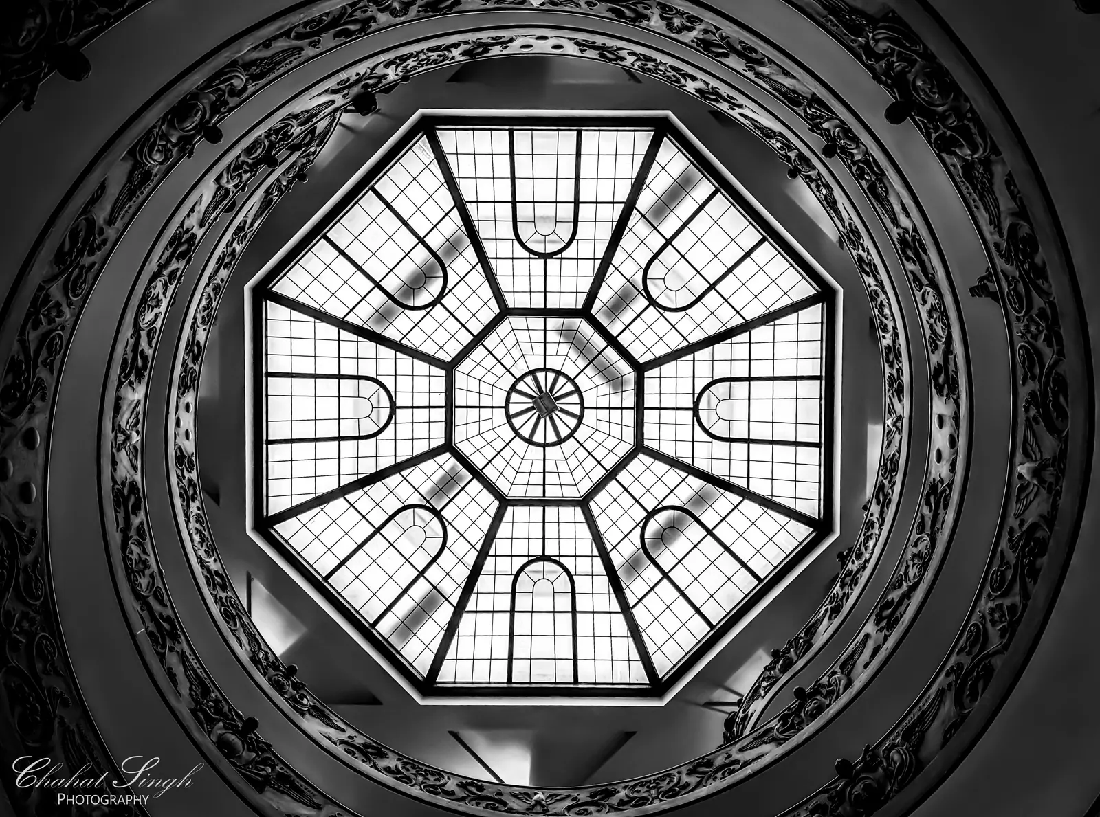 Looking straight up into an octagonal glass skylight above a spiral staircase, in black and white.